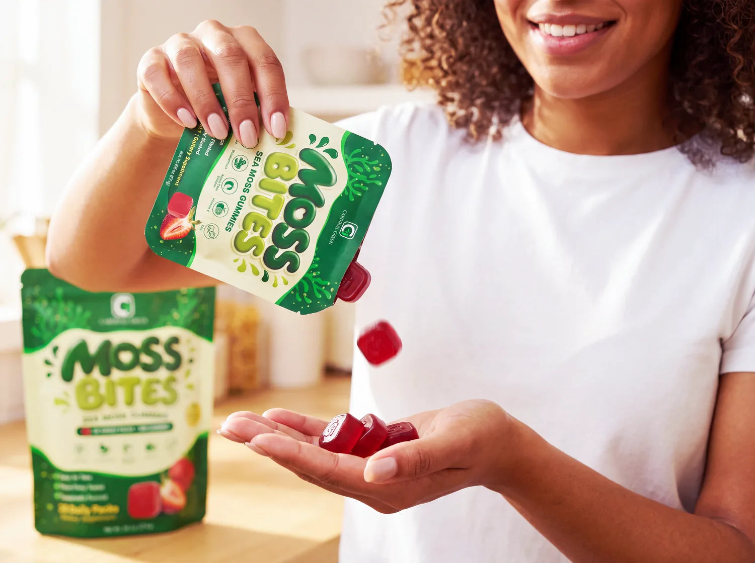 A hand tearing open a Moss Bites daily pack and pouring the strawberry gummies into an open palm, with the 28-pack bag standing on the counter beside it